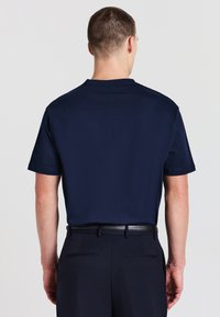 Uomo che indossa una t-shirt a maniche corte blu navy infilata in pantaloni blu navy abbinati con una cintura nera, visto da dietro su uno sfondo semplice.