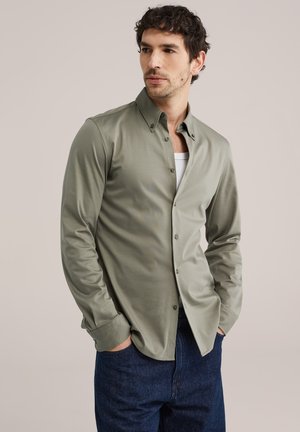 Mężczyzna w jasnooliwkowej koszuli z guzikami nałożonej na białą podkoszulkę, w ciemnoniebieskich dżinsach, stojący na jednolitym tle.