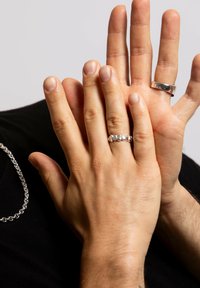 To hænder sammenflettet, den ene prydet med en sølvring og en tekstureret ring. En sølv kædehalssmykke er synlig i baggrunden.