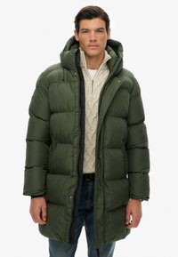 Hombre vestido con un abrigo de invierno acolchado verde oscuro con capucha, suéter de punto beige y jeans azules, de pie contra un fondo blanco.