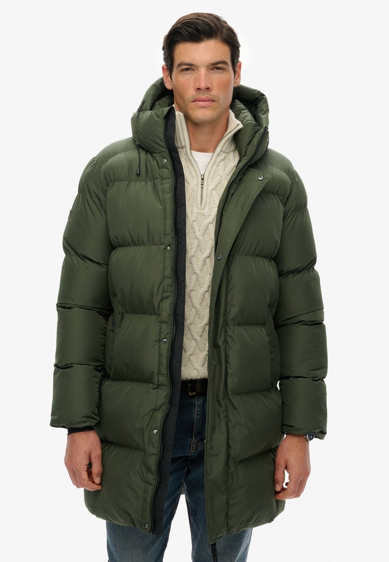 Hombre vestido con un abrigo de invierno acolchado verde oscuro con capucha, suéter de punto beige y jeans azules, de pie contra un fondo blanco.
