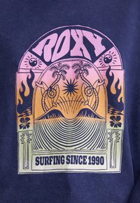 Le tissu texturé bleu marine présente un graphisme multicolore avec des palmiers, des vagues et le texte « ROXY » ; mettant en avant le surf depuis 1990.