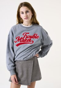 Sweatshirt gris avec le texte rouge "Trouble Maker", associé à une jupe plissée à carreaux noirs et blancs. Tissu doux et coupe décontractée.