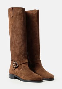 O pereche de botine înalte din suede maro, cu tocuri negre joase și curele decorative cu catarame din argint, expuse pe un fundal alb simplu.