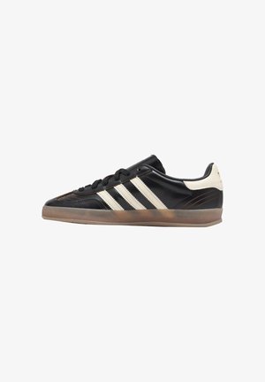 Sneaker nera low-top con strisce bianche, suola in gomma e linguetta sul tallone color crema mostrata di profilo laterale.
