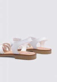 Sandalias blancas con parte superior texturizada, diseño abierto, correas ajustables y suela marrón claro, con una marca rosa en la plantilla.