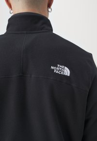 Černá fleecová bunda s vysokým límcem. Obsahuje bílé vyšívané logo na levém rameni a viditelné detaily šití.