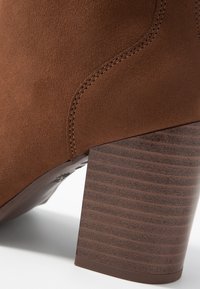 Brązowy zamszowy botek za kostkę z teksturowanym, szerokim obcasem i zakrzywionymi przeszyciami. Obcas posiada rowki dla lepszej przyczepności.