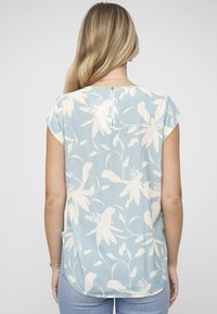 Blouse bleu clair à motifs floraux blancs ; manches courtes à capuchon, ourlet arrondi et détail zippé au dos. Texture de tissu douce.