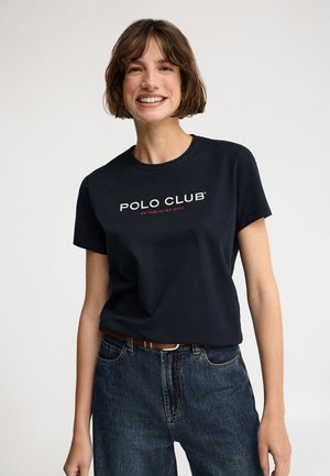 Mujer sonriente con cabello corto y castaño, viste una camiseta negra con la inscripción "POLO CLUB" metida dentro de unos vaqueros azules con cinturón marrón, sobre un fondo liso.