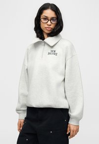 Ανοιχτό γκρι φούτερ με φερμουάρ στο ένα τέταρτο και ψηλό γιακά, με το λογότυπο "NEW BALANCE" σε σκούρο μπλε. Άνετο ύφασμα με χαλαρή εφαρμογή.