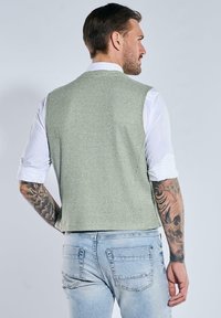 Mann mit hochgerollten weißen Hemdsärmeln und tätowierten Armen, der eine hellgrüne strukturierte Weste und eine hellblaue Jeans trägt, von hinten gesehen.