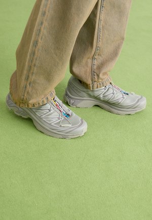 Baskets sportives gris clair avec tige en mesh, semelle extérieure texturée et lacets élastiques. Portées avec un jean beige délavé et ample sur une surface verte.