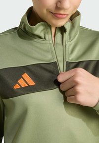Zelená bunda s vysokým golierom, s tmavozeleným horizontálnym prúžkom a oranžovým logom Adidas. Hladká textúra, zips na krku.
