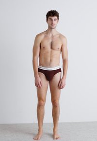 Barna rövid alsónadrág fehér derékkal, amelyen a "Calvin Klein" logó szerepel. A modell mezítláb áll egy világos háttér előtt.