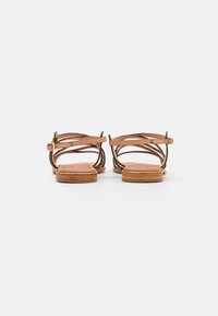 Bruine leren sandaal met kruisbanden, een platte zool, verstelbare enkelband en kruissteeksdetails. Soepele textuur en minimalistisch ontwerp.