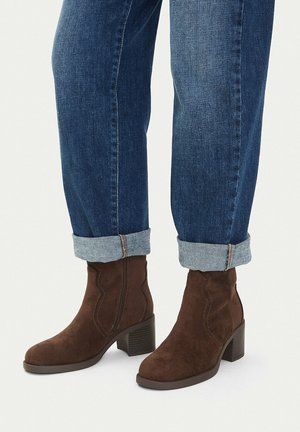 Person wearing cuffed blue jeans and brown suede ankle boots with block heels standing on a light surface.