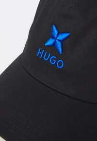 Fekete sapka szőtt anyagból, ívelt vízszintes résszel, kék hímzett márkalogóval és "HUGO" felirattal az elején.