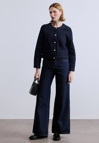 Σκούρο τζιν μπουφάν με ασημένια κουμπιά και δύο μπροστινές τσέπες, συνδυασμένο με φαρδιά σκούρα τζιν παντελόνια. Το μοντέλο κρατάει μια μικρή μαύρη τσάντα.