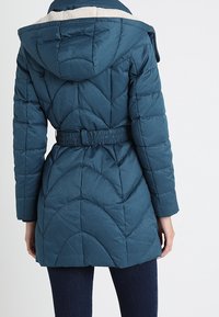 Kølig, quilted puffer-jakke i teal med hætte, udstyret med et blødt indre foder, bælte i taljen og buede søm på ryggen.