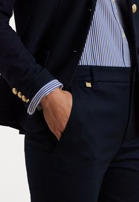 Granatowa marynarka z złotymi guzikami i mankietami niebieskiej koszuli w paski. Spodnie dopasowane z złotym akcentem na kieszeni. Gładkie, teksturowane tkaniny.