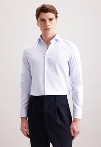 Camicia azzurra chiaro con colletto classico, maniche lunghe e tessuto morbido. Abbinata a pantaloni scuri a vita alta.