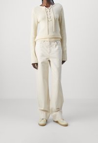 Pull en tricot beige avec détail à lacets au cou, associé à un pantalon large beige clair et des chaussures slip-on beige. Texture lisse tout au long.