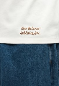 Λευκή ραφή πουκάμισου με καφέ κέντημα «New Balance Athletics, Inc.» πάνω από μπλε τζιν.