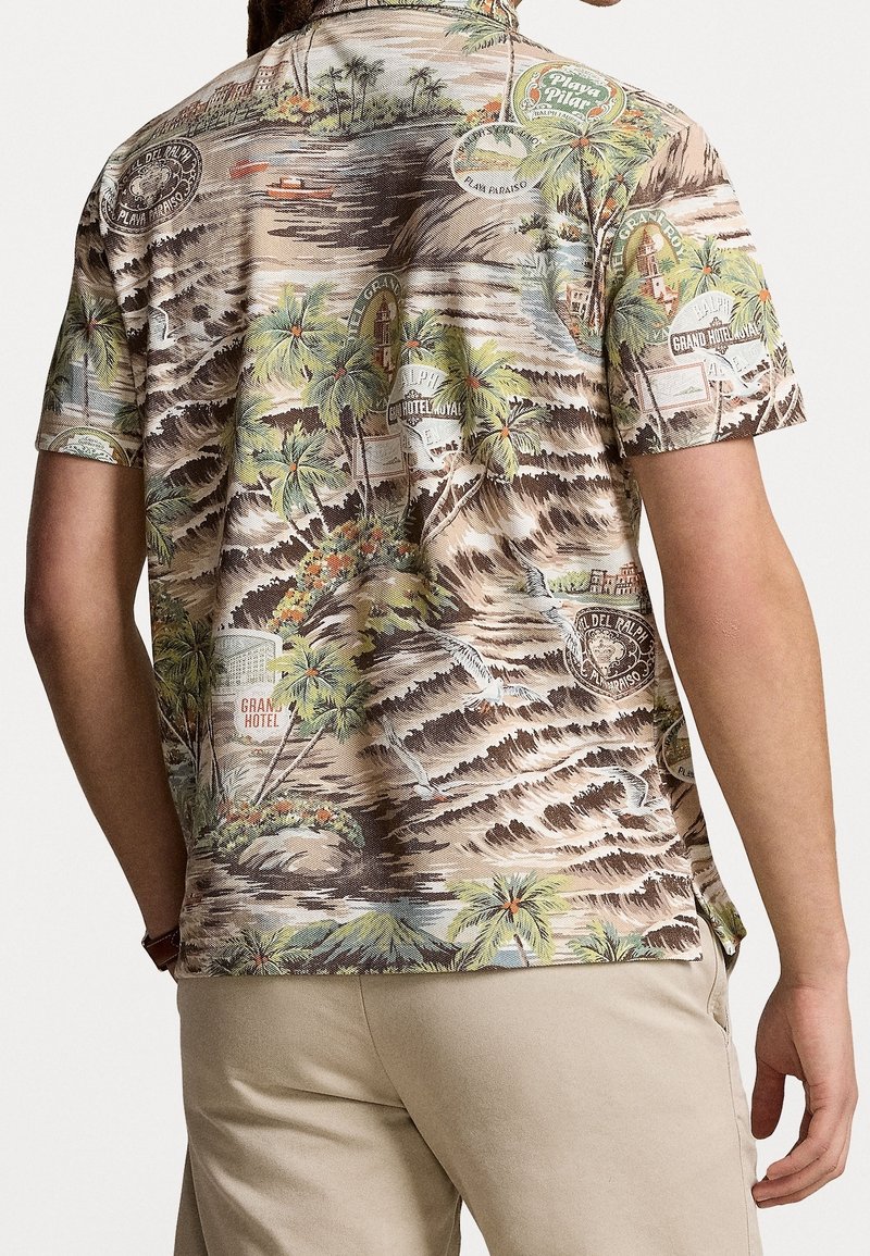 Homme portant une chemise à manches courtes à imprimé tropical avec des palmiers, des vagues, des enseignes d'hôtel et des oiseaux, associée à un pantalon beige, vu de dos.