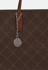 Brązowa torebka w wzory z tan skórzaną rączką i srebrnym łańcuchem trzymającym okrągły metalowy znacznik grawerowany napisem "Tamaris".