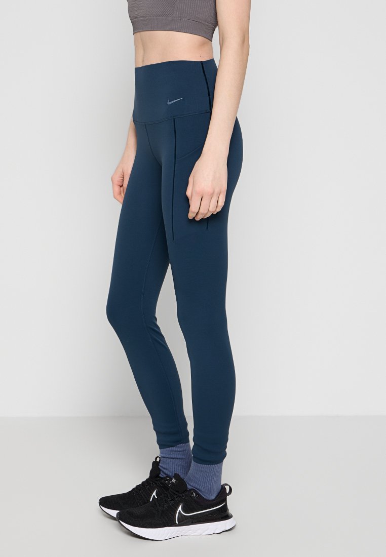Leggings de tiro alto en azul marino, hechos de una tela suave y elástica, con un logo sutil y un bolsillo lateral. Combinados con zapatillas deportivas negras.