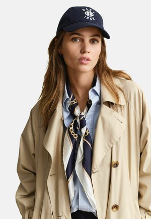 Jeune femme portant un trench-coat beige, une chemise bleue à boutons, une écharpe à motifs bleu marine et blanc, et une casquette de baseball bleu marine avec des lettres blanches.