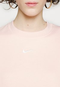 Světle růžová mikina Nike s kulatým výstřihem a vyšívaným bílým logem na hrudi, vyrobená z měkké, texturované látky.