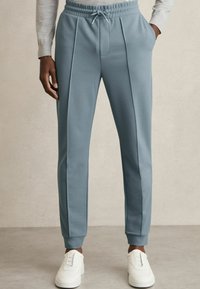 Pantaloni de trening de culoare albastru deschis, dintr-un material neted, cu un cordon la talie, având două pliuri frontale și manșete ribbate, asortați cu adidași albi.
