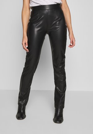 Kvinde iført tætsiddende sorte læderbukser med høj talje og sorte ankelstøvler, stående mod en ensfarvet lysegrå baggrund.