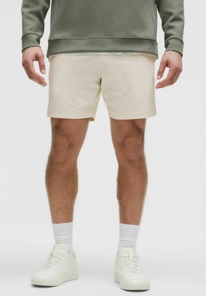 Hombre con pantalones cortos beige claro, calcetines blancos, zapatillas blancas y una sudadera verde, de pie frente a un fondo liso.