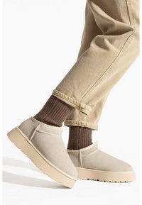 Zapatos slip-on de ante beige con una suela gruesa beige, combinados con calcetines marrones de canalé y pantalones claros remangados.
