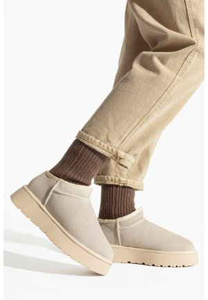 Scarpe slip-on in suede beige con suola spessa beige, abbinate a calze a coste marroni e pantaloni chiari arrotolati.