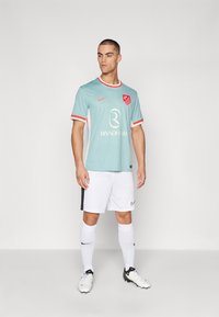 Camisa de fútbol azul claro con detalles rojos, pantalones cortos blancos y logo negro de Nike. La camisa presenta un emblema del club y texto en el centro.