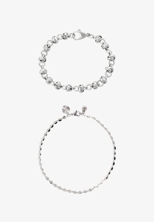 Dwie srebrne bransoletki na białym tle; jedna masywna z skręconymi ogniwami i zapięciem typu karabińczyk, druga cienka z małymi zawieszkami w kształcie dysków i zapięciem typu karabińczyk.