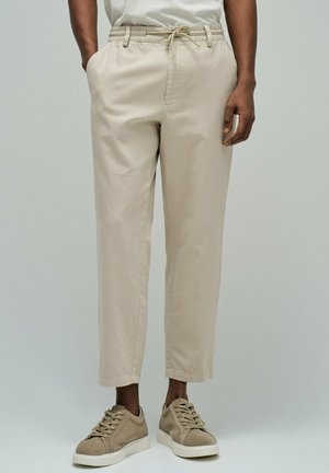 Homme portant un pantalon beige à cordon et des baskets beige assorties, debout avec les mains détendues le long du corps.