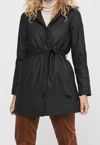 Svart regnrock med knäppning fram, insvängd midja och huva. Slätt, vattentätt material med långa ärmar och sidofickor.