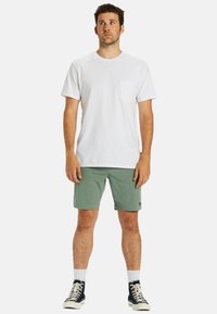 T-shirt en coton blanc avec poche, shorts verts et baskets noires avec semelles blanches. Coupe décontractée, design simple, sans motifs ni graphiques visibles.