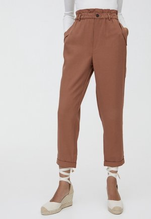 Pantalon paperbag marron taille haute avec ourlets retroussés, porté avec des espadrilles compensées blanches à lacets à la cheville et un haut blanc à manches longues rentré.