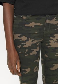 Kamouflage-printade jeans i gröna och bruna nyanser, tillverkade av stretchmaterial, med femficksdesign och dekorativt stygn.