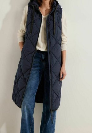 Femme portant un long gilet matelassé sans manches bleu marine avec une capuche par-dessus un haut crème à manches longues et un jean bleu sur un fond uni.