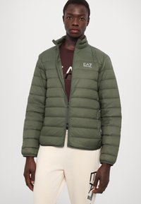 Green puffer jacket with horizontal quilted design, high collar, zip closure, and logo on the chest, paired with light-colored pants.