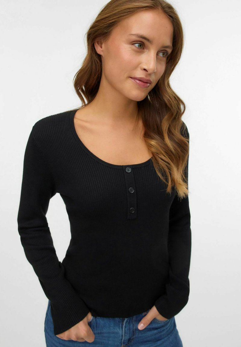 Giovane donna con capelli mossi, che indossa una maglietta nera a maniche lunghe a coste con tre bottoni e jeans blu, mani in tasca, che guarda a destra.