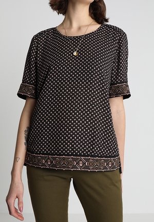 Vrouw draagt een zwart patroon blouse met korte mouwen en decoratieve randen, gecombineerd met olijfgroene broek, afgebeeld van schouders tot heupen.