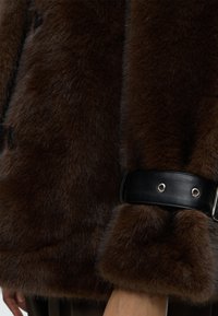 Σκούρο καφέ χνουδωτό μανίκι με φαρδιά μαύρη δερμάτινη λωρίδα και ασημένιες οπές γύρω από τον καρπό, φορεμένο στον βραχίονα ενός ατόμου.